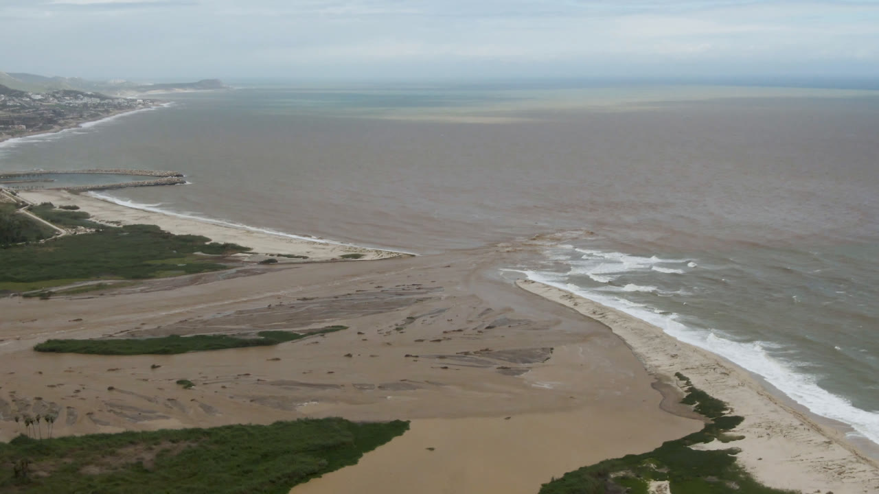 A second aerial frame of San Jose del Cabo shoreline shows surf crossing sand banks, green vegetation at the lower edge, open water beyond, and no visible people.