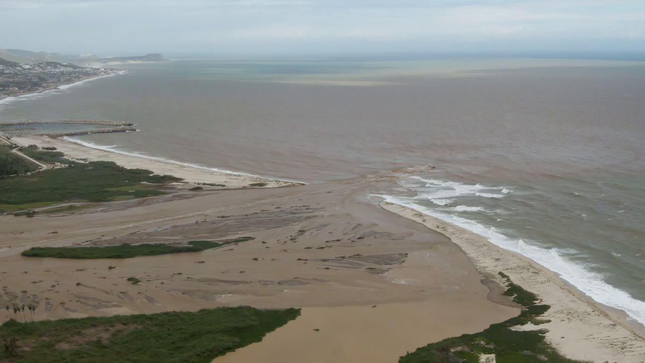 Drone-like aerial view of a beach, delta water, waves, and distant shoreline.