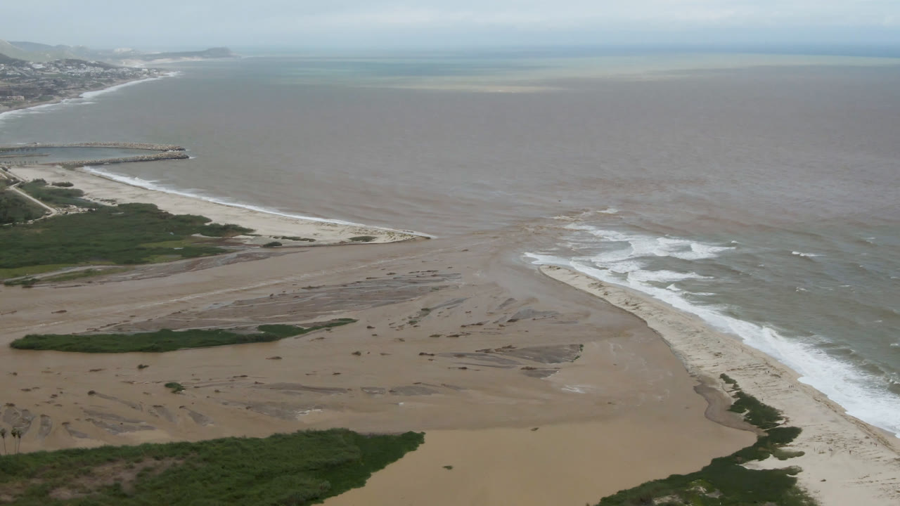 Aerial frame over San Jose del Cabo showing tan sand banks, white surf, green vegetation in the foreground, distant coastline, and no visible people.