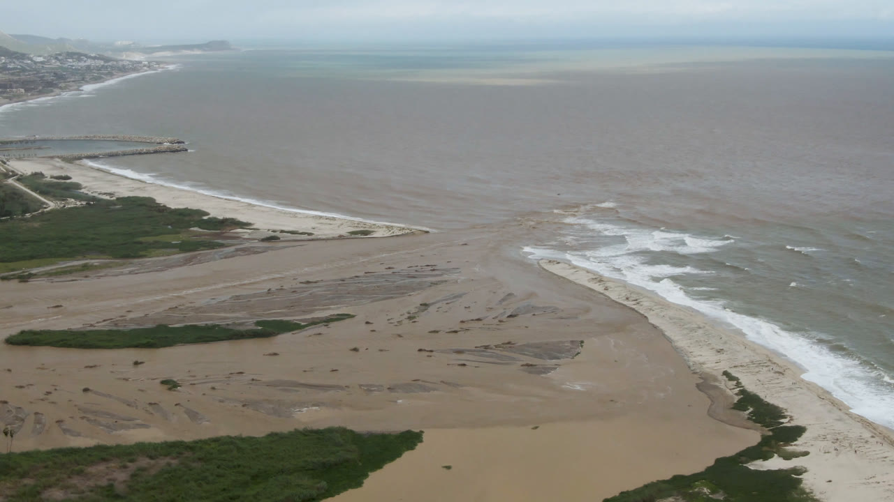 Aerial frame over San Jose del Cabo with surf bands curling across shallow brown-green water, broad sand flats below, and no visible people.