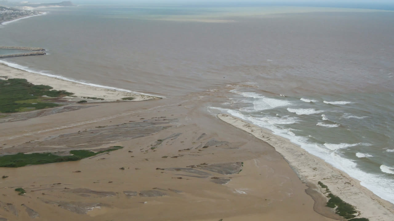 Aerial frame over San Jose del Cabo showing a wide tan beach, white surf along the shore, green vegetation at the lower edge, brown nearshore water, and no visible people.