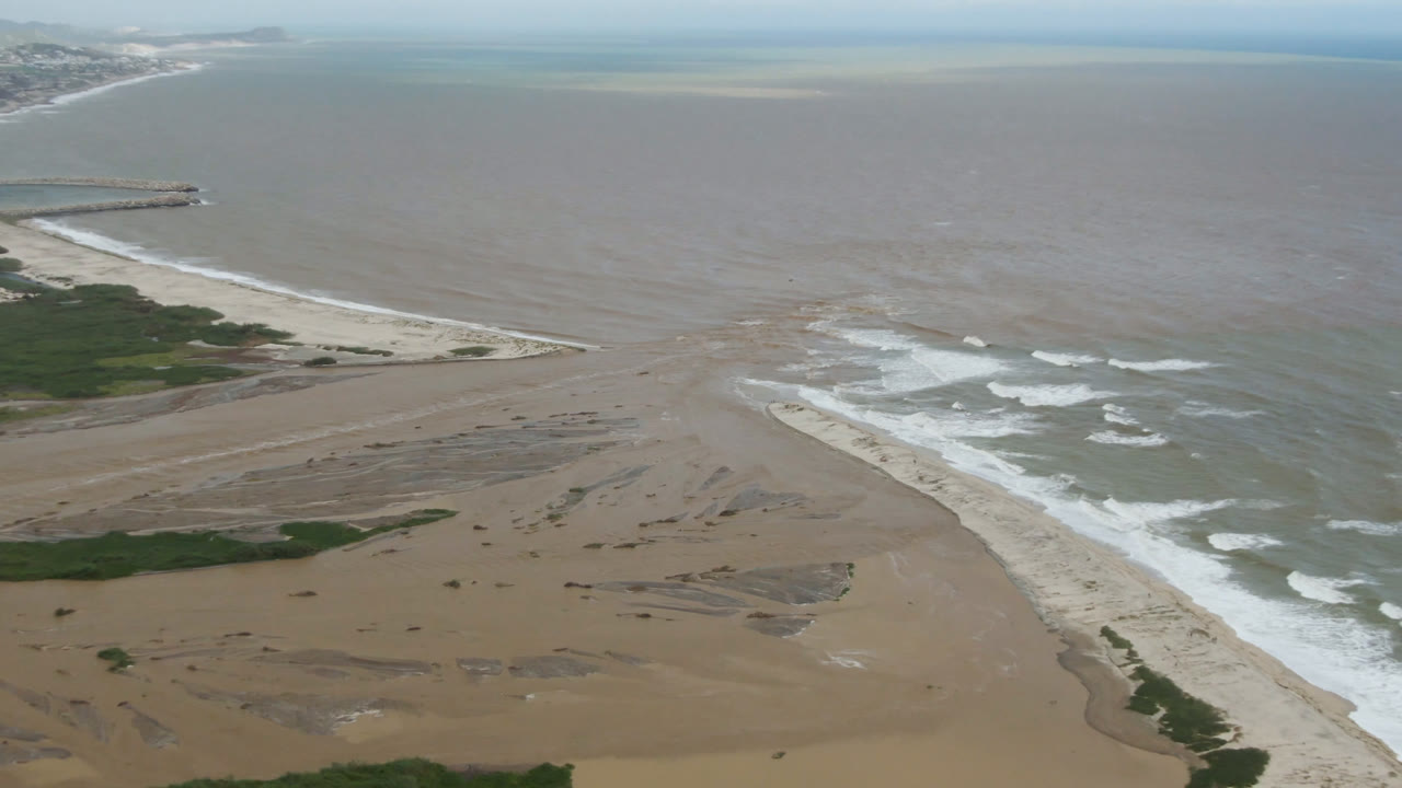 Aerial view in San Jose del Cabo with the sandy shoreline running diagonally, surf breaking in white rows, muted blue and brown water beyond, and no visible people.