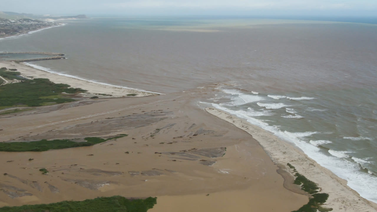 Aerial shoreline with broad beach, surf, vegetation patches, and hazy coast