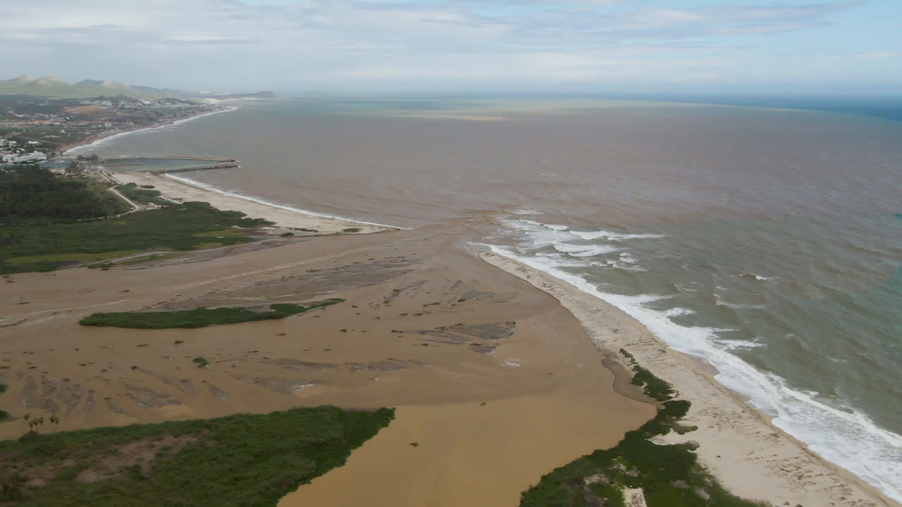 Aerial view over San Jose del Cabo showing a sandy river mouth entering the ocean, green riverbank vegetation, distant shoreline development, cloudy sky, and no visible people.