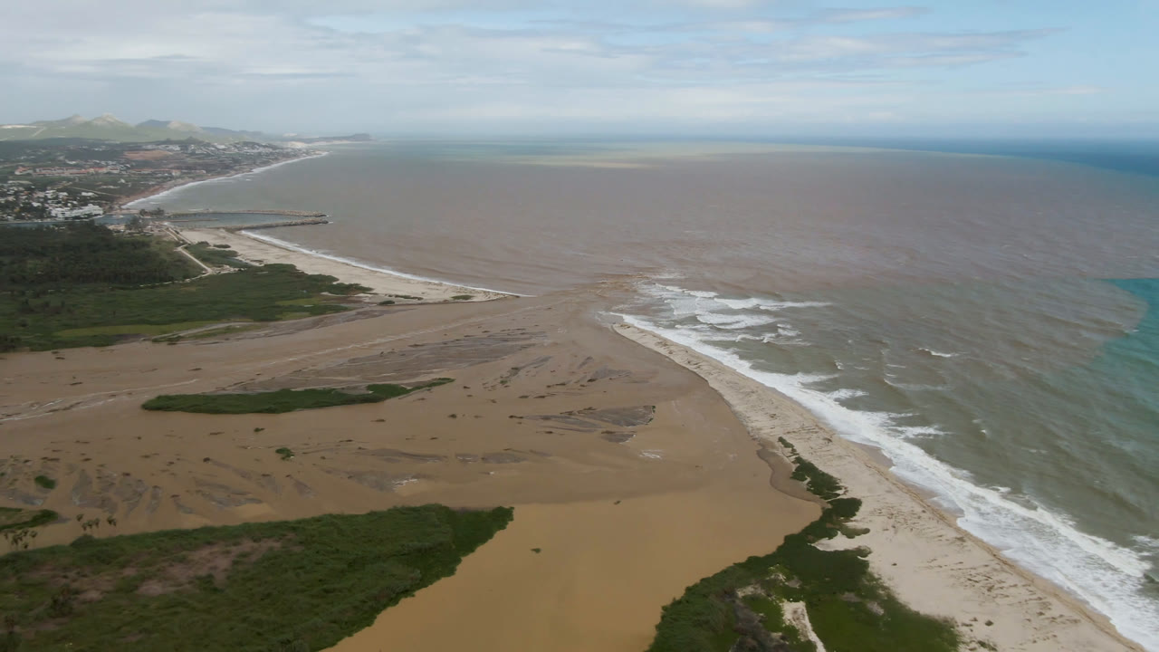 Wide aerial frame in San Jose del Cabo with brown river water spreading across the beach edge, blue ocean water to the right, white surf, distant hills, and no visible people.