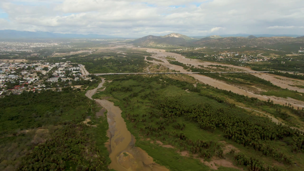 Opening aerial frame over San Jose del Cabo showing a muddy river through dense green vegetation, a town edge at left, distant mountains, clouded daylight, and no visible people.
