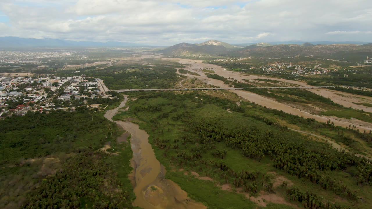 Second aerial frame in San Jose del Cabo with the muddy river crossing the valley, clustered buildings near the left midground, roads, mountains, and no visible people.