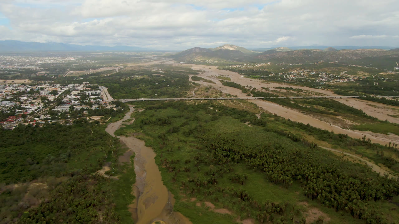 Wide aerial view of a muddy river through a green valley with mountains beyond.