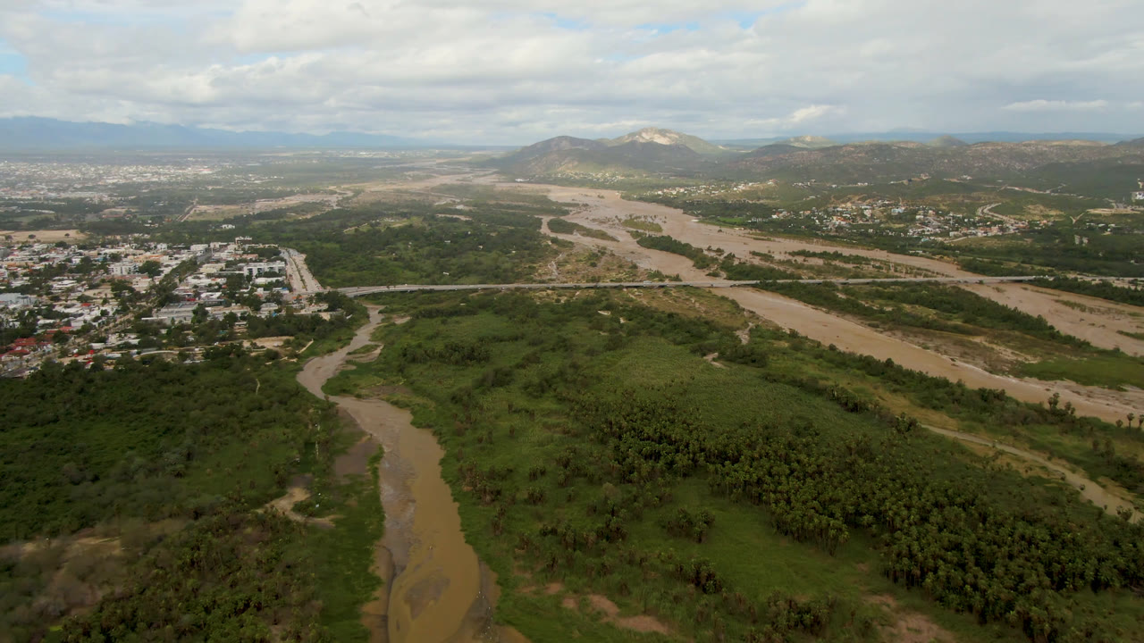 Aerial view in San Jose del Cabo with the muddy river running through green valley growth, a road crossing the scene, buildings in the midground, distant hills, and no visible people.