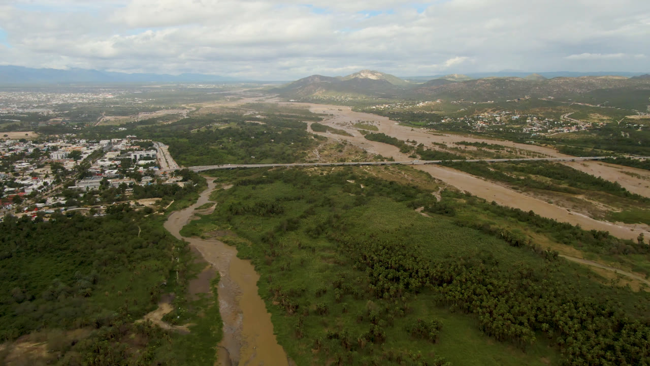 Opening aerial frame over San Jose del Cabo, Baja California Sur, showing a muddy river, dense green vegetation, nearby town blocks, distant hills, clouded sky, and no visible people.