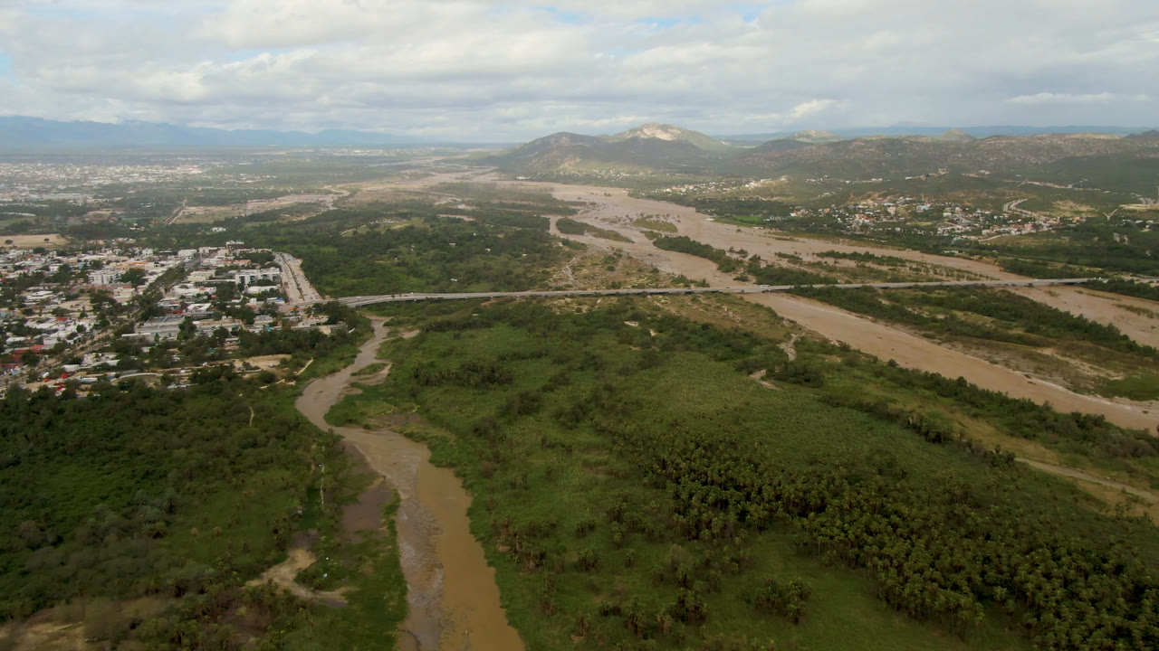 Second aerial frame over San Jose del Cabo with the river channel stretching diagonally through green brush, a developed town area to the left, hills behind it, and no visible people.