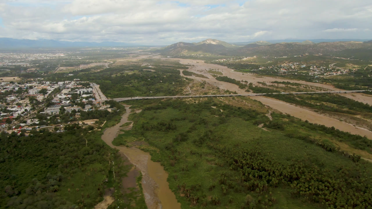 Wide aerial frame from San Jose del Cabo, Mexico, showing a muddy river curving through dense green vegetation with buildings, a bridge, distant hills, clouded sky, and no visible people.