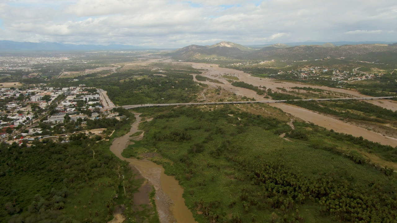 Aerial frame from San Jose del Cabo, Mexico, with the brown river running diagonally past green brush, a road bridge, clustered low buildings, hazy hills, heavy clouds, and no visible people.