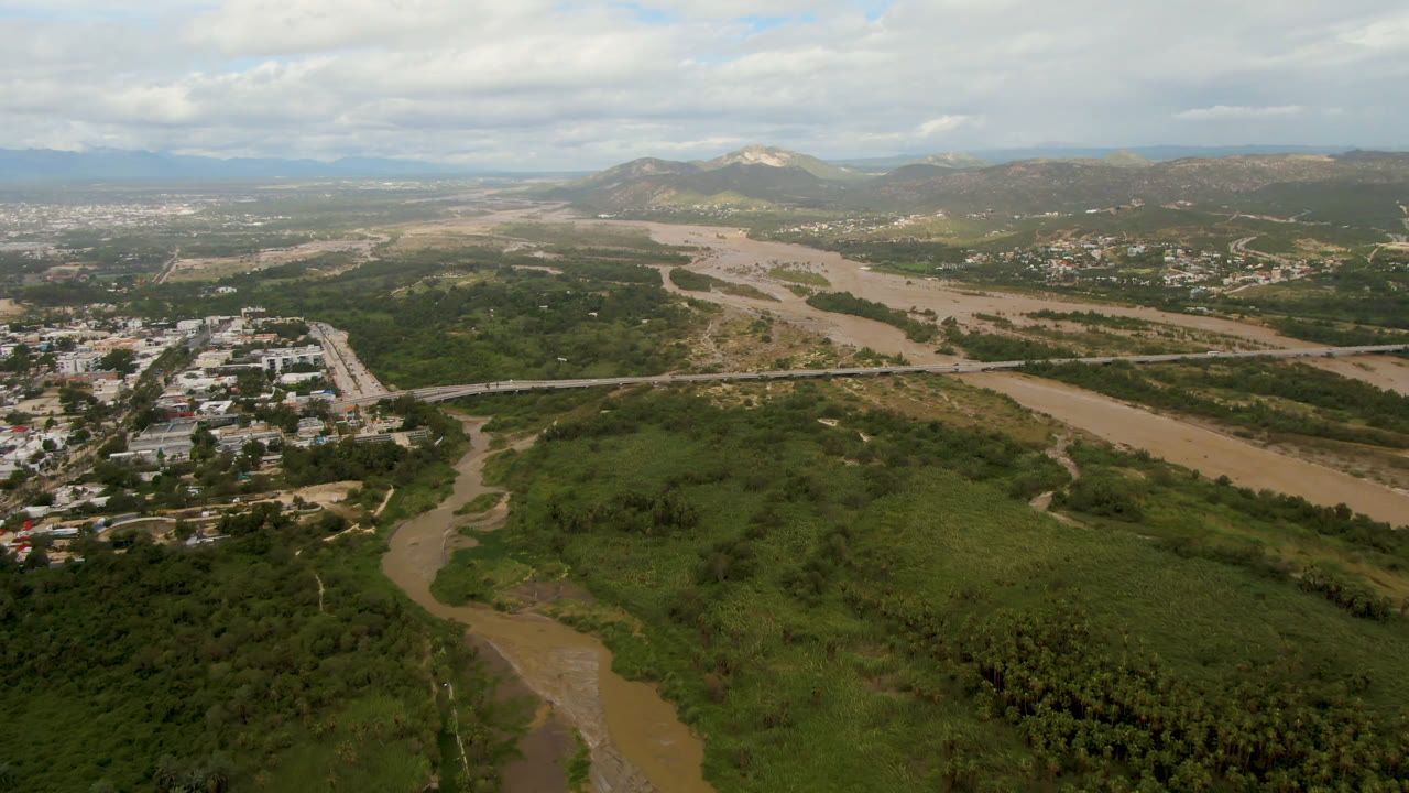 Wide aerial frame over San Jose del Cabo, Baja California Sur, with a muddy river, dense green valley, roads, distant buildings, mountains, clouded sky, and no visible people.