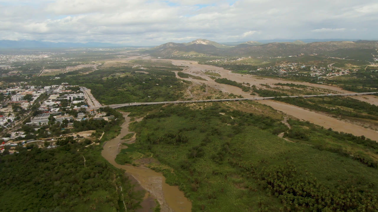 Aerial view in San Jose del Cabo showing the river bend below, a road bridge crossing the valley, clustered buildings to one side, low mountains, overcast daylight, and no visible people.