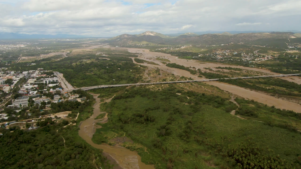 Opening aerial frame over San Jose del Cabo shows a muddy river curving through dense green vegetation, light buildings at left, distant hills, cloudy sky, and no visible people.