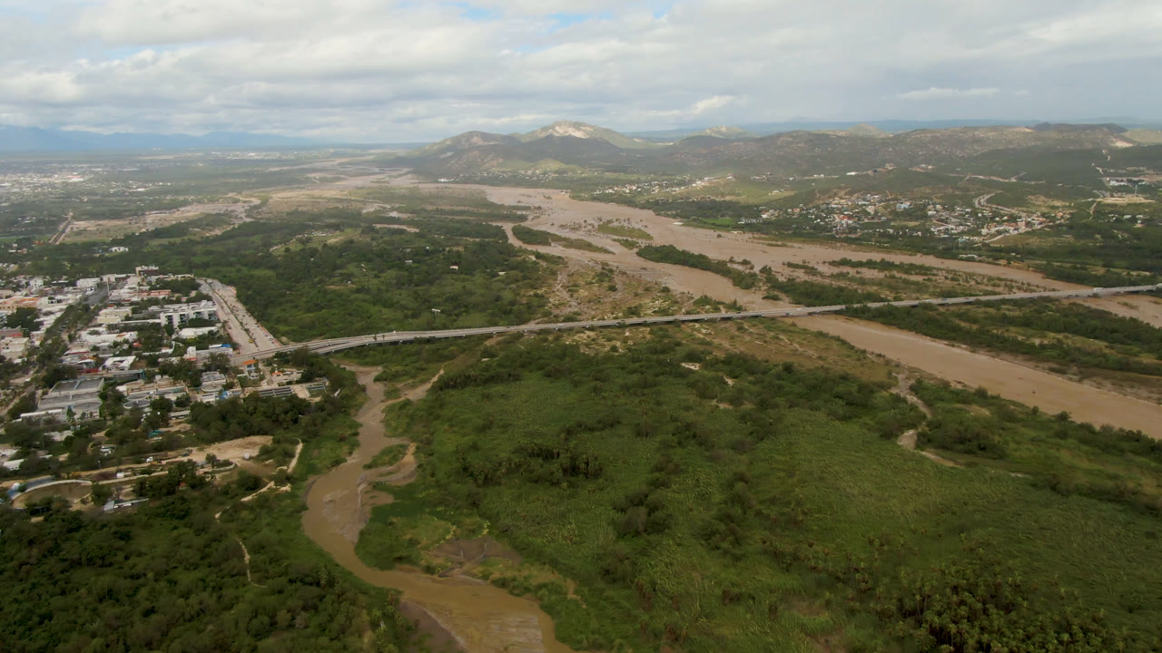 Second aerial frame in San Jose del Cabo centers the river and green floodplain, with a road crossing the midground, buildings near the left edge, hills beyond, and no visible people.