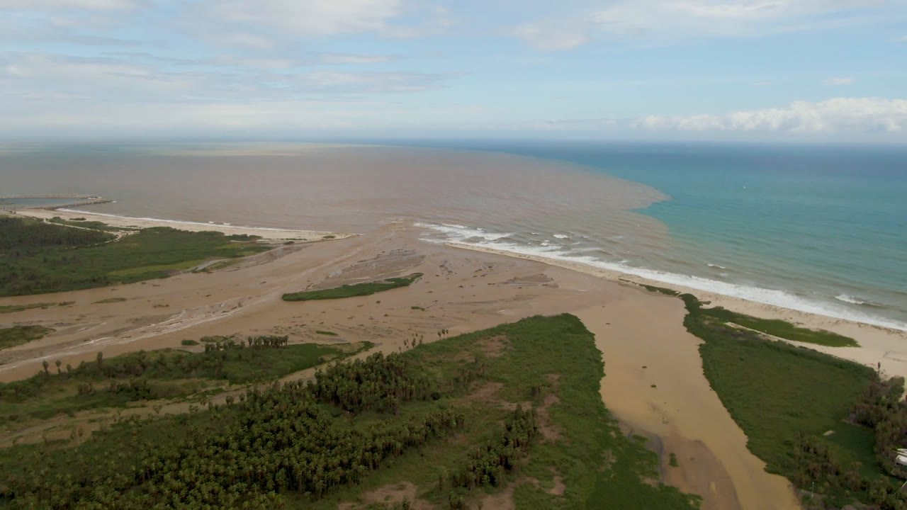 Aerial frame in San Jose del Cabo, Baja California Sur, with the river mouth centered, pale surf breaking along the sand, green coastal vegetation in the lower frame, and no visible people.