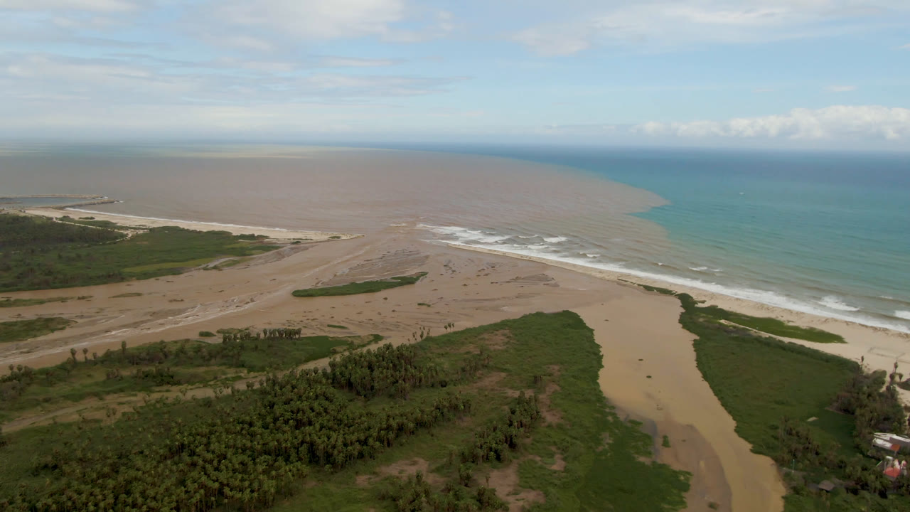 Aerial view over San Jose del Cabo with green vegetation in the foreground, a sandy river channel reaching the beach, brown water entering turquoise ocean, and no visible people.