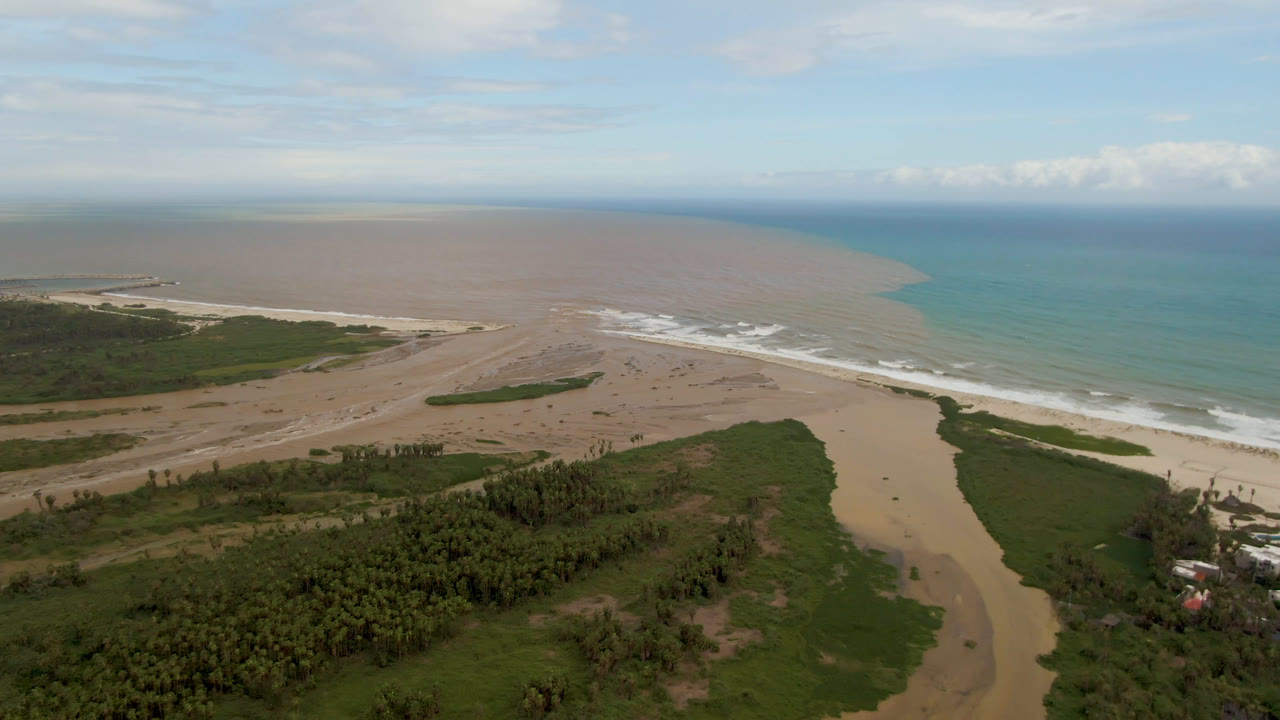 Aerial view in San Jose del Cabo with the river channel centered below, pale sandbars stretching toward the surf, green vegetation in the lower frame, turquoise ocean at right, and no visible people.