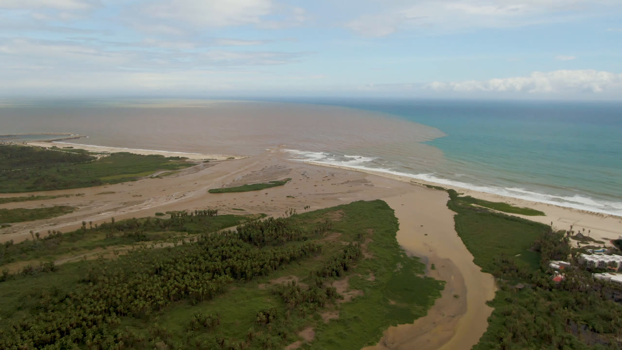 Aerial view of a sandy river mouth meeting the ocean beside green coastal vegetation.