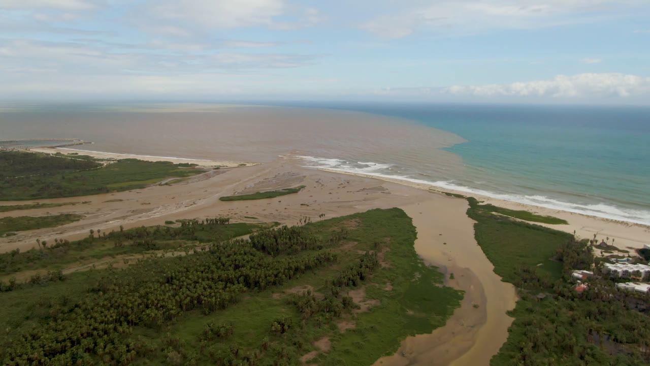 Aerial view in San Jose del Cabo with a pale sand delta centered below, darker river water running toward the surf, green shoreline vegetation, open ocean, clouds, and no visible people.