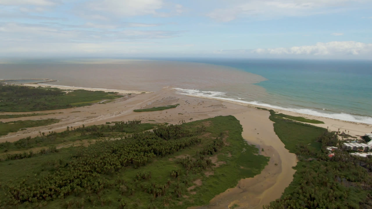 Aerial view in San Jose del Cabo, Baja California Sur, with a river channel cutting through green vegetation toward surf and a brown plume beside blue ocean, no visible people.
