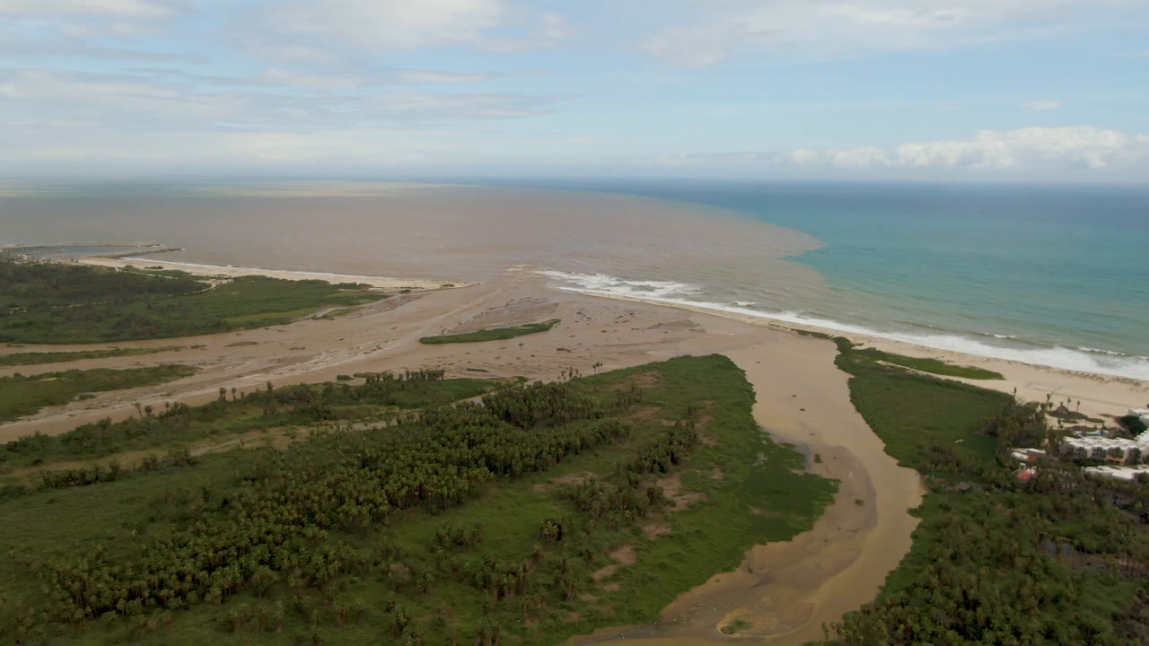 Wide aerial frame over San Jose del Cabo showing the river mouth below, pale beach and white surf across the middle, and blue ocean fading into a muddy offshore plume, no visible people.