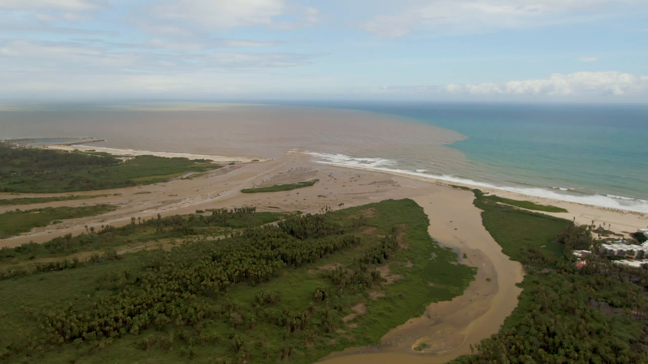 Drone view of beach, river delta, green shoreline, and two-toned ocean water.