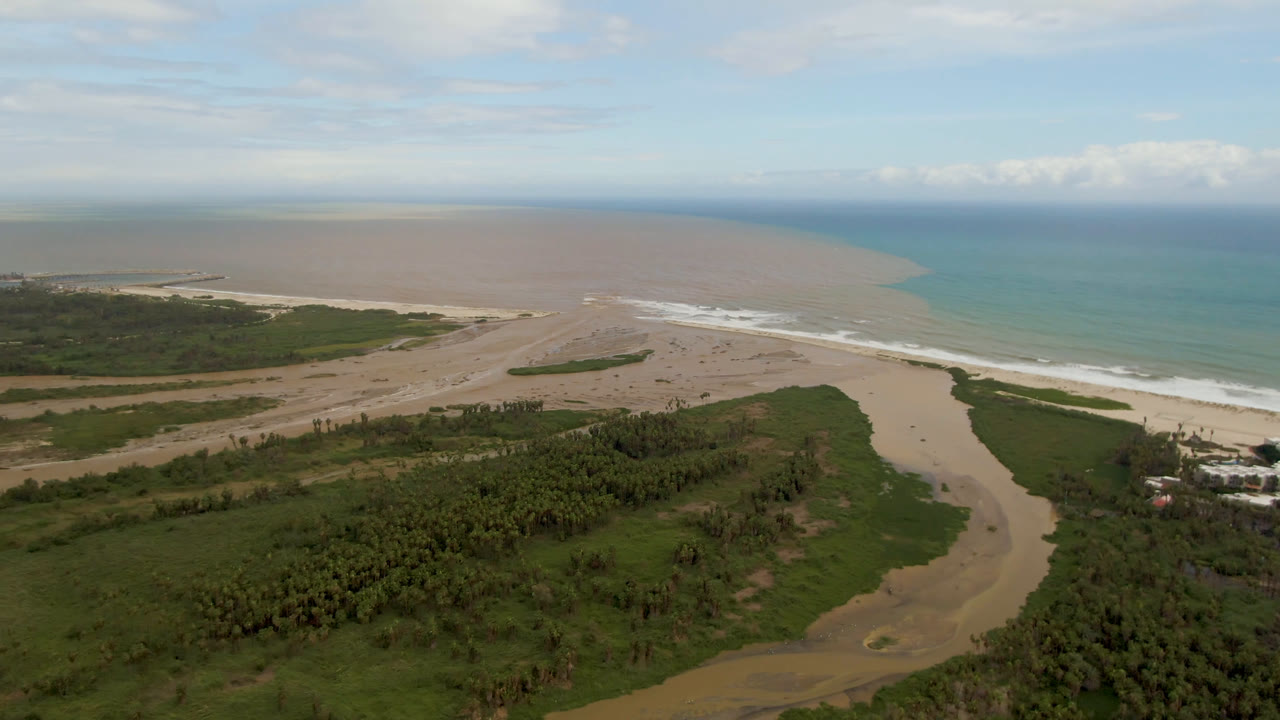 Wide aerial frame over San Jose del Cabo showing a tan river channel through green coastal vegetation, pale beach, blue-green ocean, and no visible people.