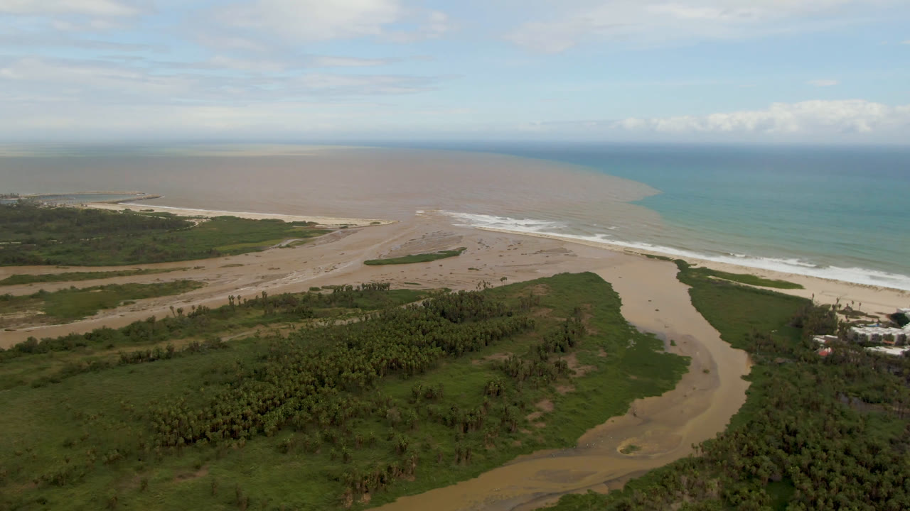 Aerial view in San Jose del Cabo with the river channel centered below, surf breaking along the beach, blue ocean to the right, and no visible people.