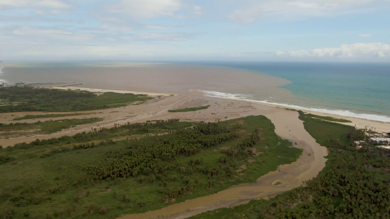 Aerial view of a winding river, green vegetation, sand beach, surf, and ocean.