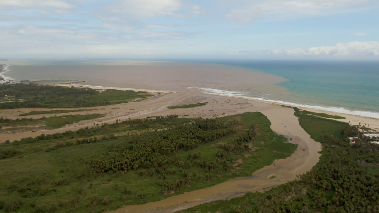 Aerial frame in San Jose del Cabo with the river channel centered below, dense green vegetation in front, sandy beach beyond, and the ocean horizon under light cloud cover, with no visible people.