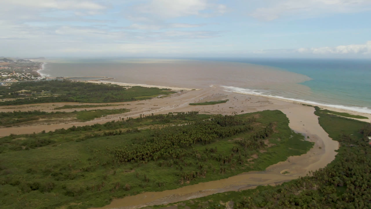 Wide aerial frame over San Jose del Cabo, Mexico, showing a brown river channel crossing green wetlands toward a sandy beach and blue ocean, with no visible people.