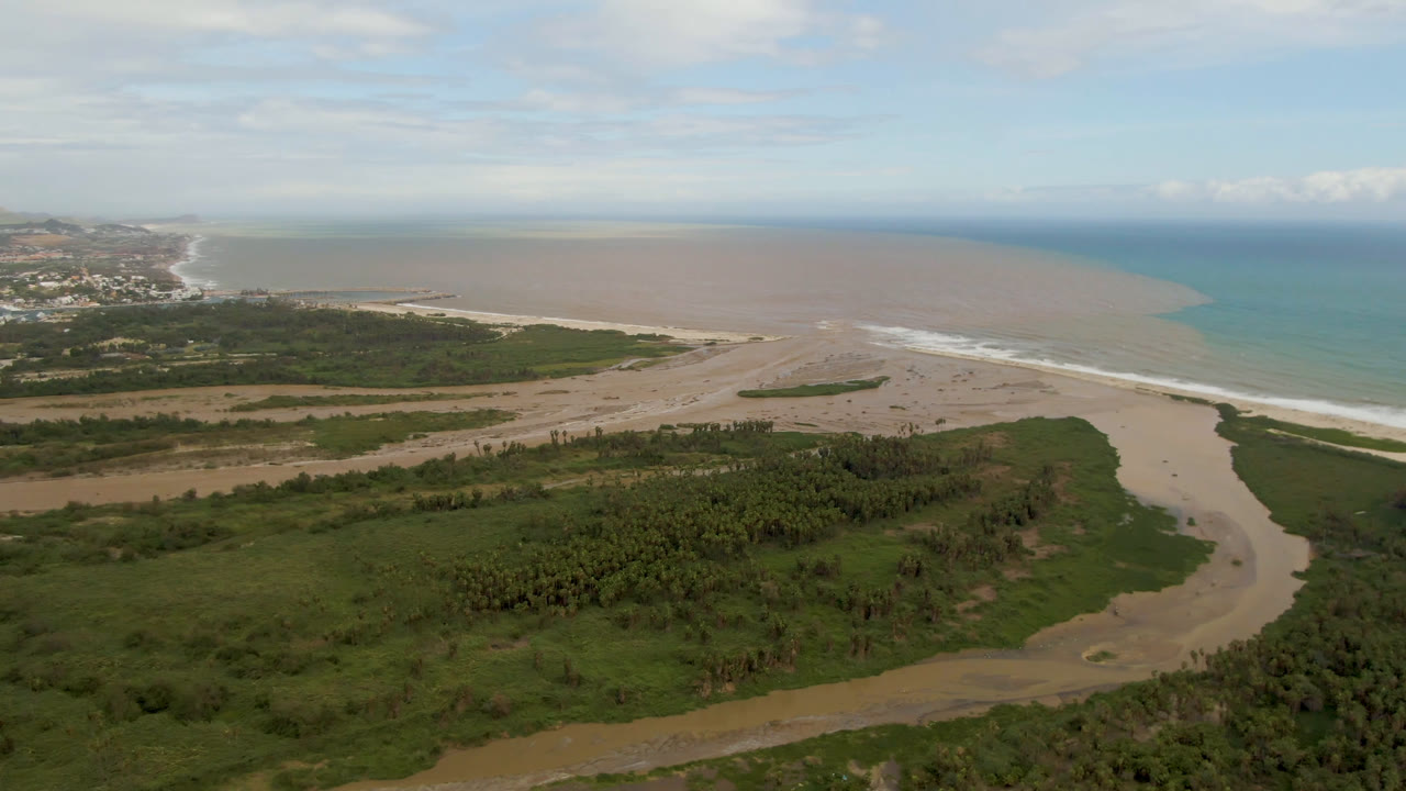 A second aerial view of San Jose del Cabo, Mexico, with the winding river more centered through green vegetation and sand before reaching the surf, with no visible people.