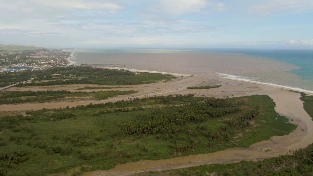 Aerial frame over San Jose del Cabo with dense green vegetation in front, sandy flats and beach in the middle, brown shallow water and blue ocean beyond, and no visible people.