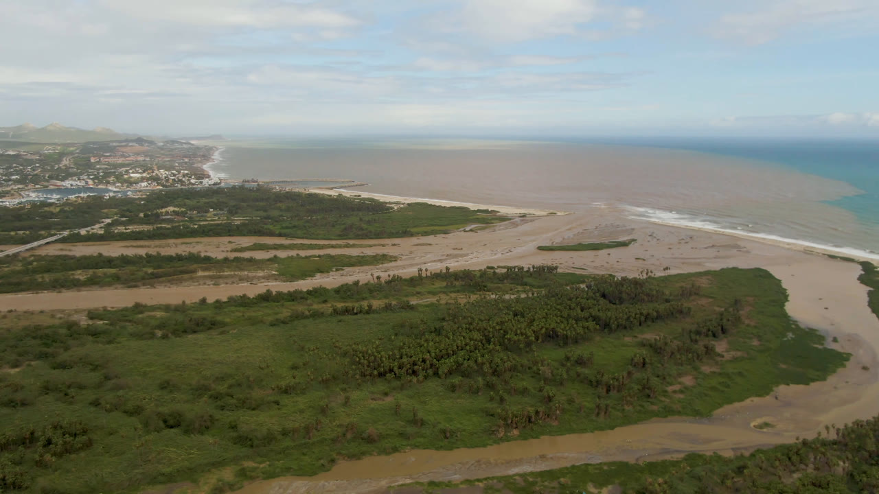 Wide aerial view in San Jose del Cabo showing foreground palms, a curving sandy channel, surf along the beach, distant buildings to the left, and no visible people.