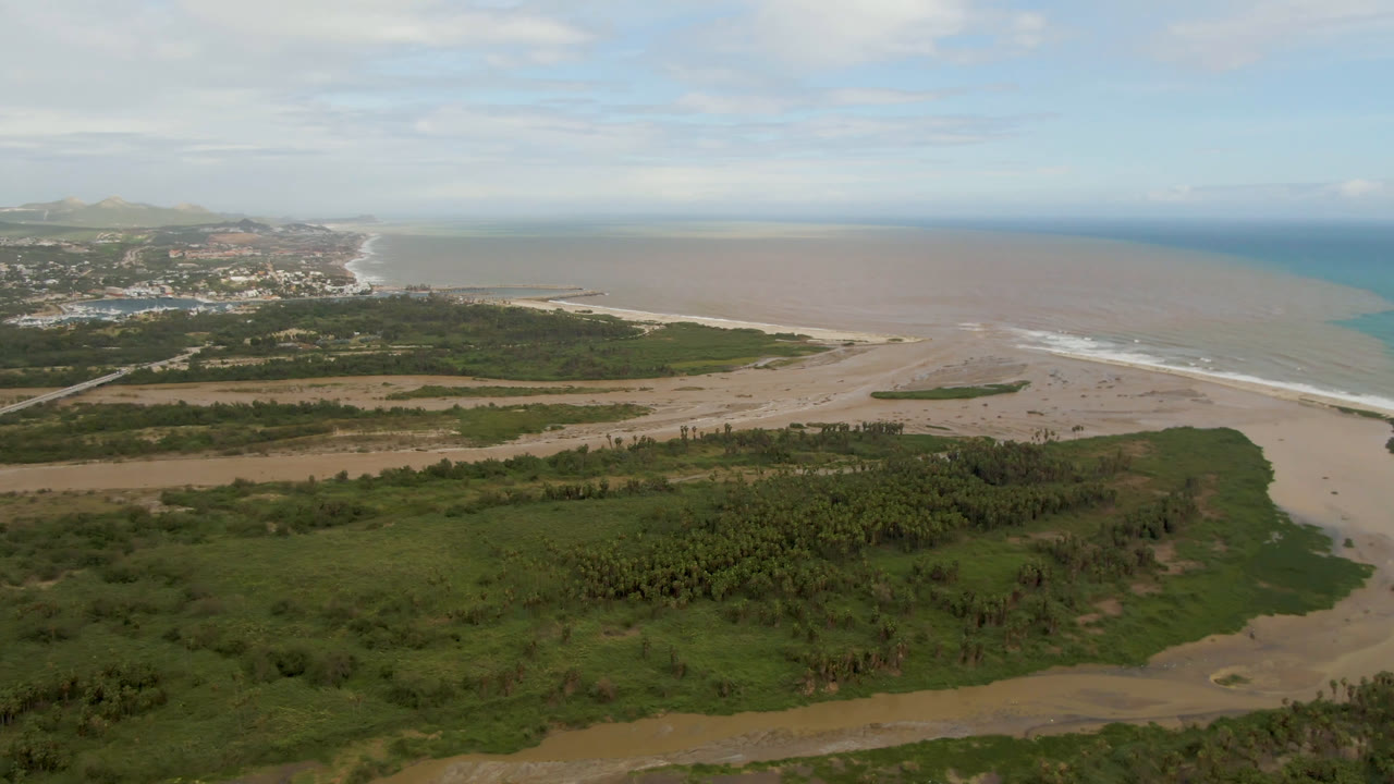 Aerial coastal landscape with beach, brown water, blue ocean, green vegetation, and distant buildings.