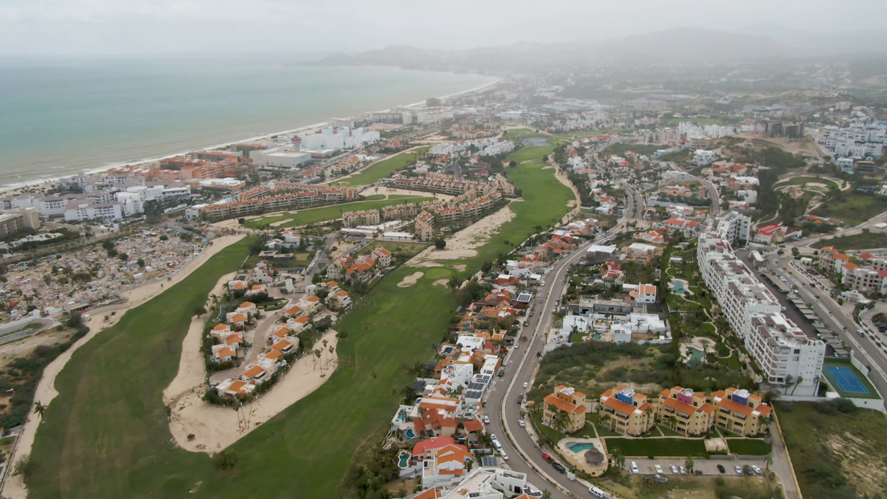 Opening frame over San Jose del Cabo shows a broad green golf course in the foreground, a curving road and red-roofed buildings through the center, and hazy beach and ocean in the distance, with no visible people.