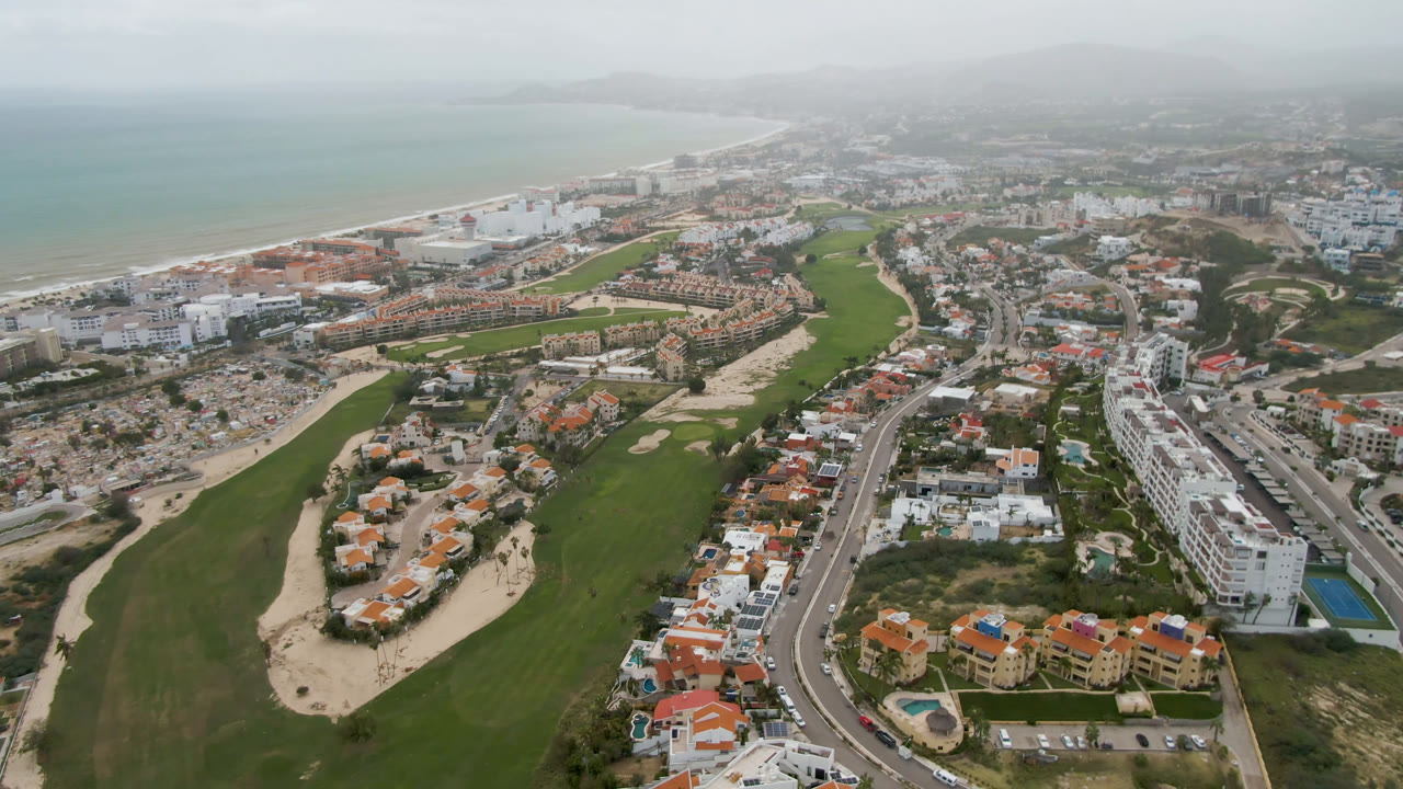 At 0:01, the San Jose del Cabo aerial keeps the golf fairway dominant at left, with dense low-rise buildings, a main road, sandy shoreline, and pale ocean beyond, with no visible people.