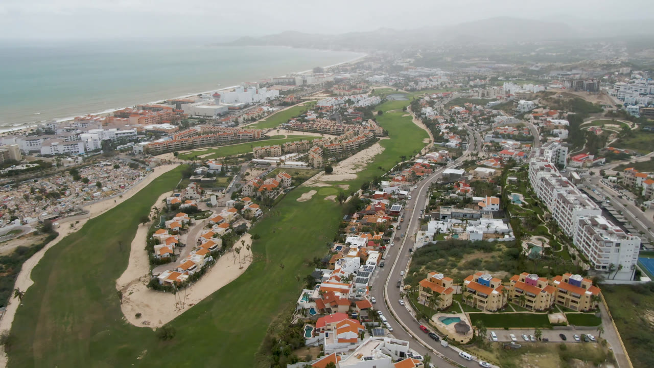Opening aerial frame over San Jose del Cabo with green fairways and sand bunkers in front, coastal road and white buildings to the right, hazy beach and ocean beyond, and no visible people.