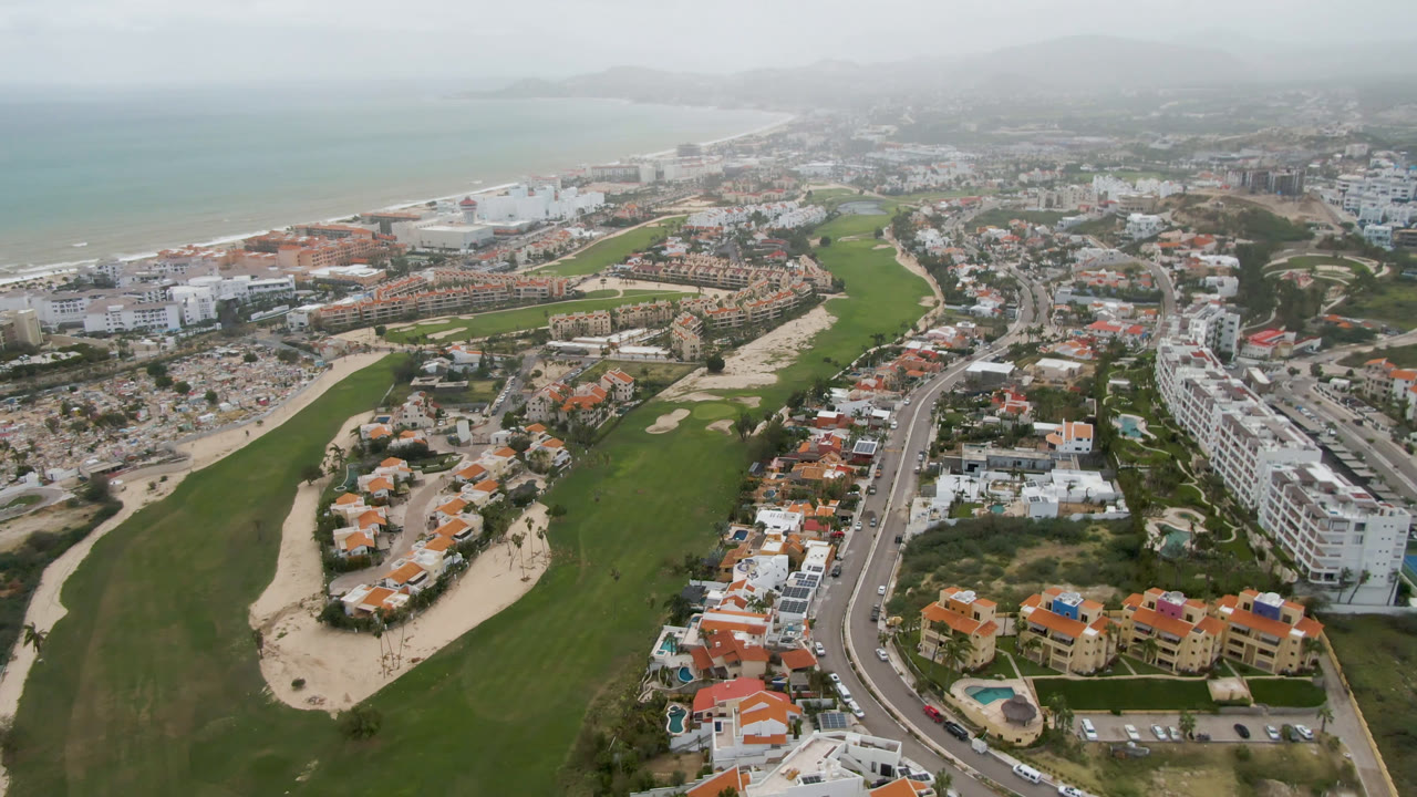 Aerial view in San Jose del Cabo with the golf course centered, a curved road and dense white buildings on the right, beach hotels near the water, hazy ocean in the distance, and no visible people.