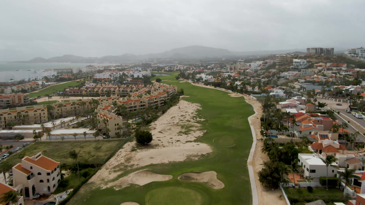 Aerial view of a golf course beside red-roofed buildings and distant water under gray clouds.