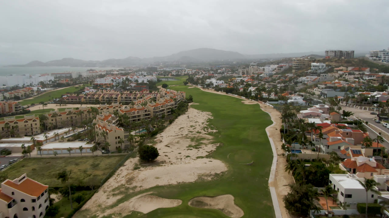 San Jose del Cabo aerial frame with a fairway running through the center, pale sandy ground on the right, homes and streets around it, distant water, and no visible people.
