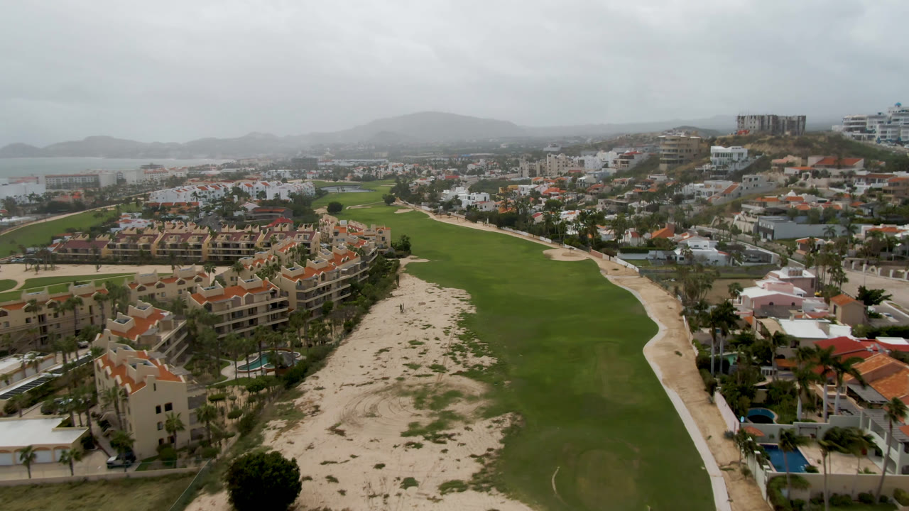 Aerial view of a golf fairway, sandy strip, coastal buildings, and distant water under gray clouds.