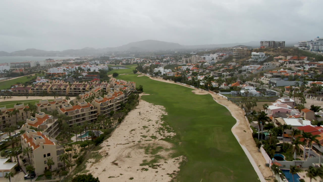 At 0:01, the San Jose del Cabo aerial view holds on fairway grass, a sandy corridor, palm-lined buildings, distant coastline, gray sky, and no visible people.