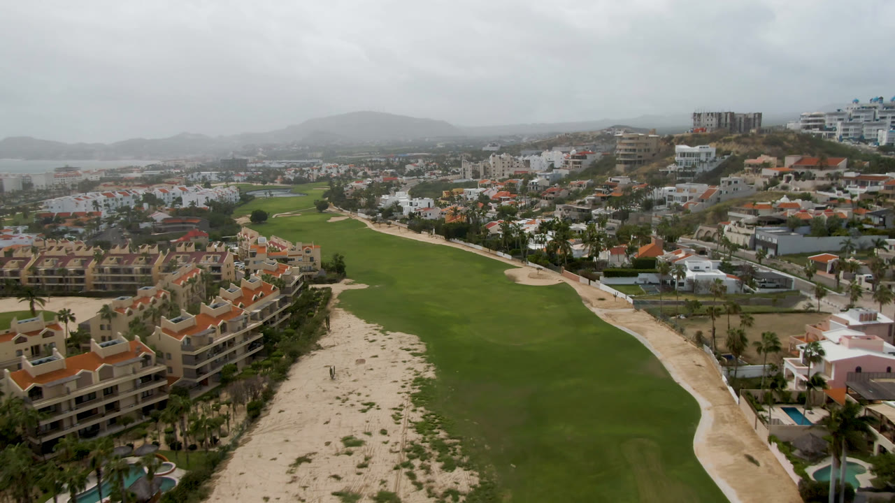 Opening aerial frame over San Jose del Cabo with a green golf course in the center, sandy paths, terracotta-roofed buildings at the lower left, a hazy bay and hills in the background, and no visible people.