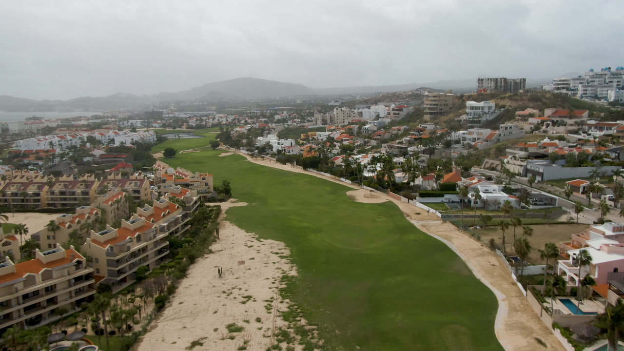 Aerial frame in San Jose del Cabo looking across central green fairways, pale sand areas, palm-lined resort buildings at the lower right, dense city blocks beyond, and no visible people.