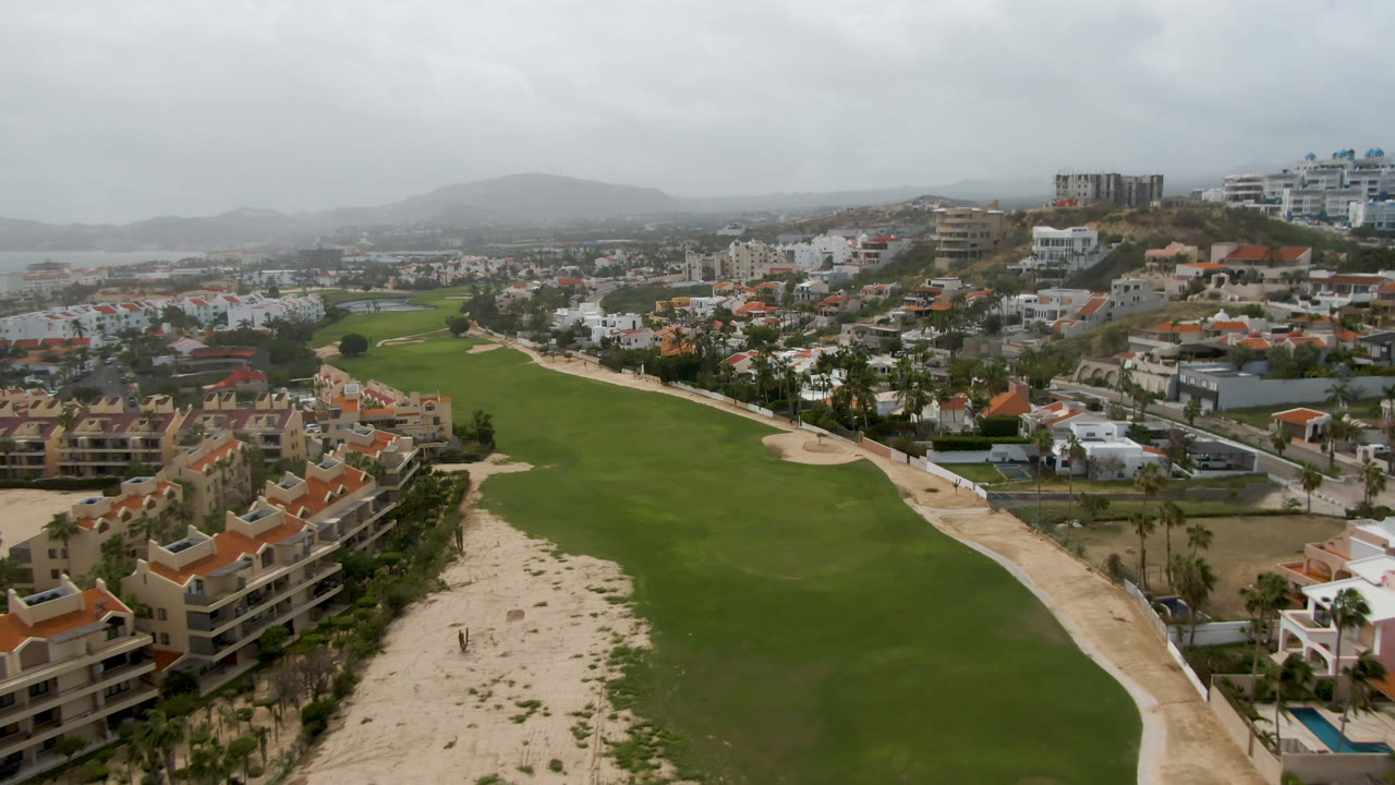 Elevated view of a golf course with sandy borders, resort buildings, city blocks, and distant hills.