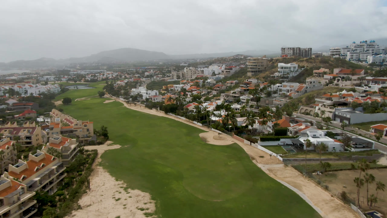 Opening aerial frame over a San Jose del Cabo golf fairway, with sand bunkers, red-roofed homes, distant hills, gray cloud cover, and no visible people.